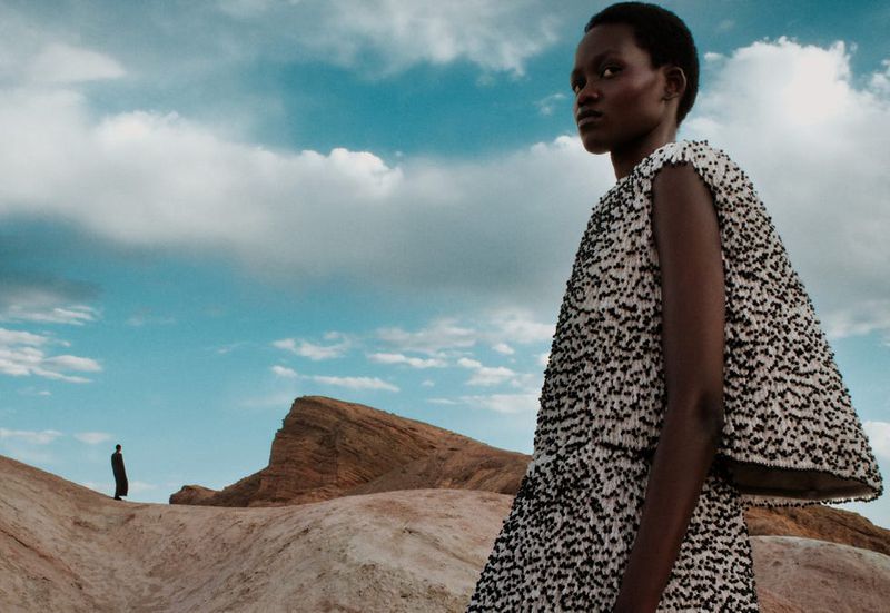 A stylish woman in a beaded dress stands against a desert backdrop with a partly cloudy sky and a distant figure in the background.