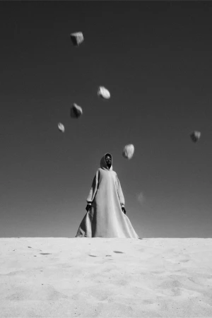 Monochrome photo of a person in a robe standing in sand with rocks falling from the sky in a desert landscape on a clear day.
