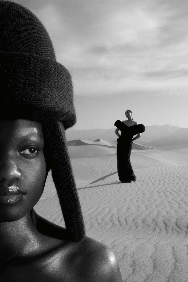 Black and white fashion photo of a model in a sculptural headpiece in the foreground with another model posing on desert sand dunes.
