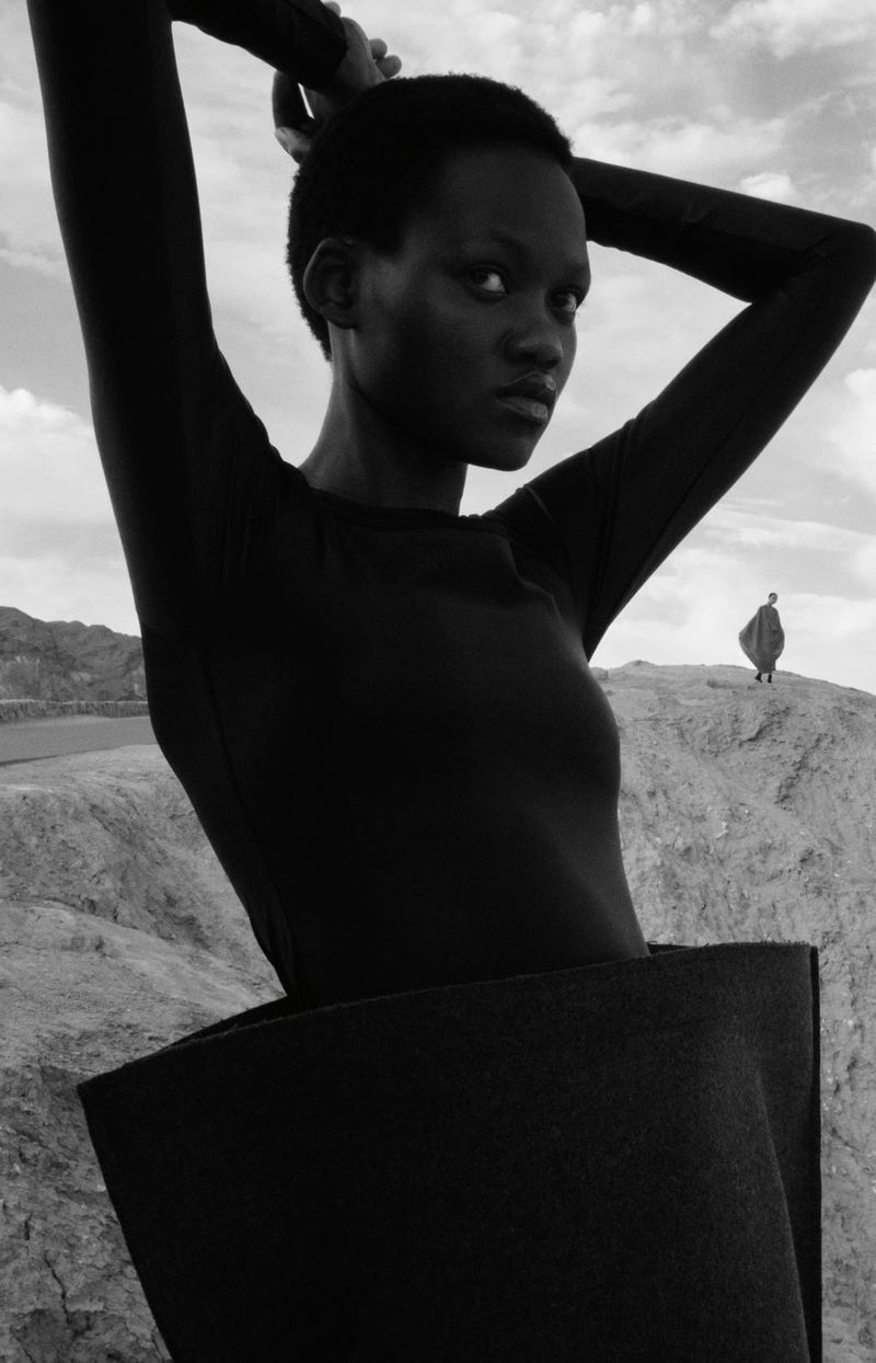 A striking monochrome fashion portrait of a dark-skinned woman with hands behind her head against an arid landscape, featuring dramatic light.