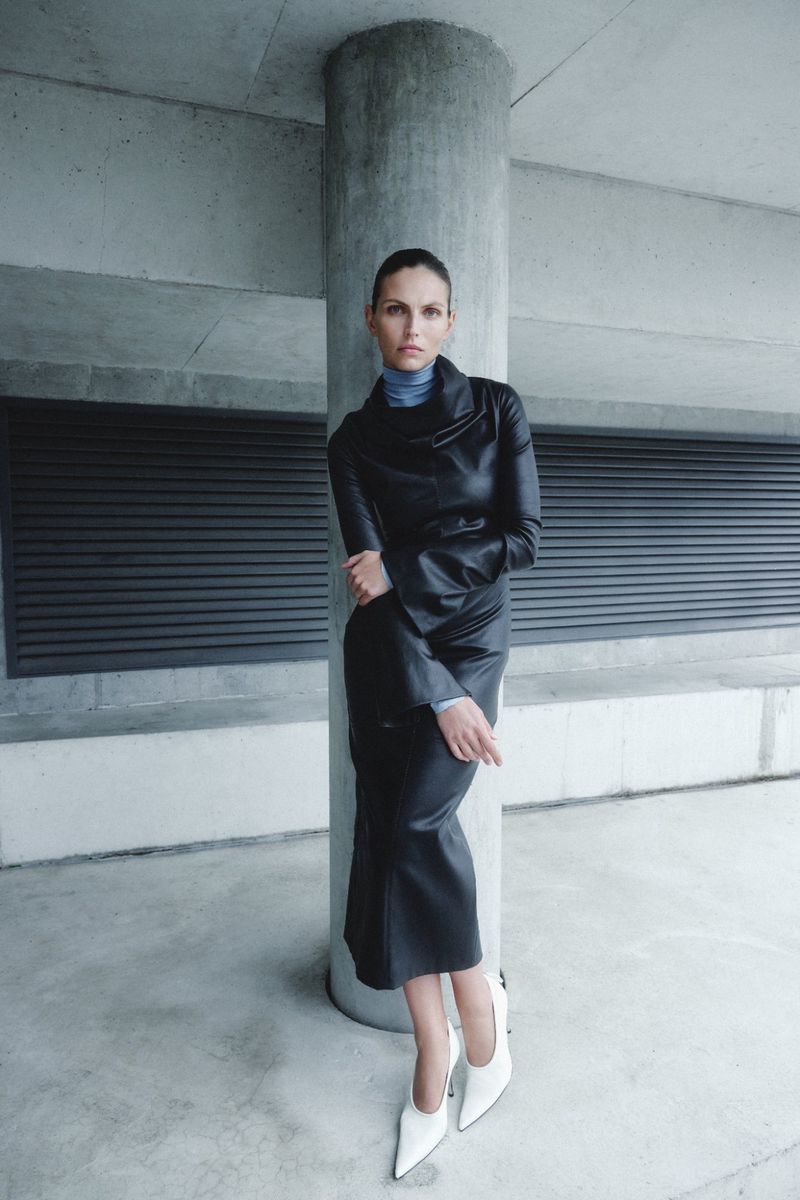 Karlina Caune leaning against a concrete pillar wearing a black leather dress, blue turtleneck, and white pointed-toe pumps.