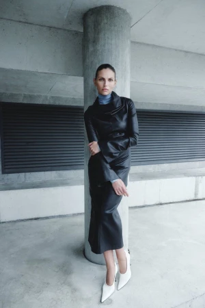 Karlina Caune leaning against a concrete pillar wearing a black leather dress, blue turtleneck, and white pointed-toe pumps.