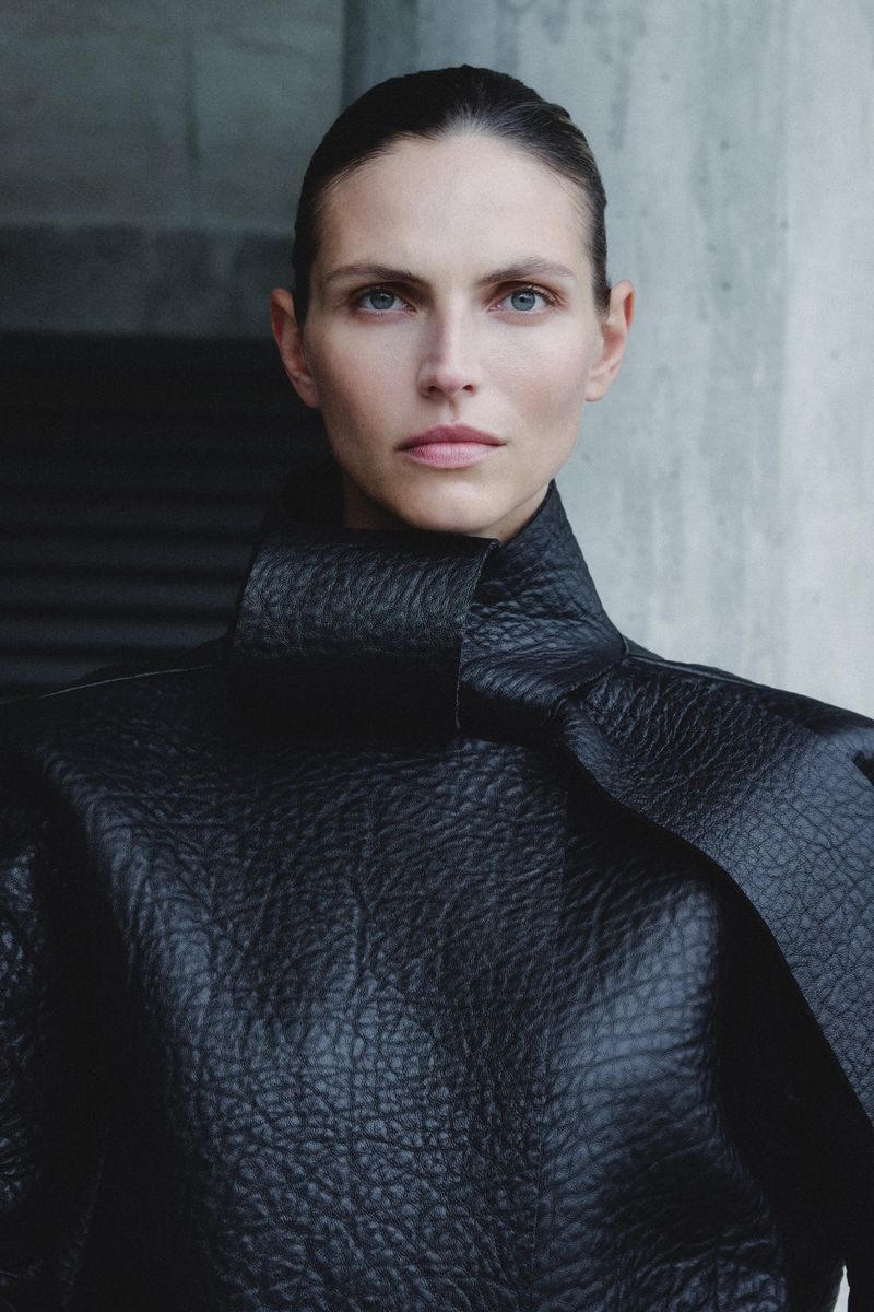 Portrait of an adult woman with neutral expression wearing a black textured top with a high collar against a concrete background.