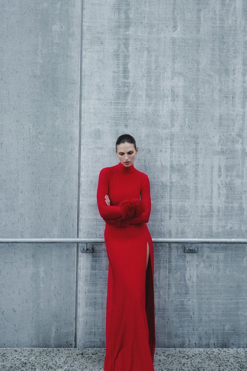 Woman in a red, long-sleeved gown poses against a concrete wall. The dress features a high neckline and thigh-high slit, fashion editorial.