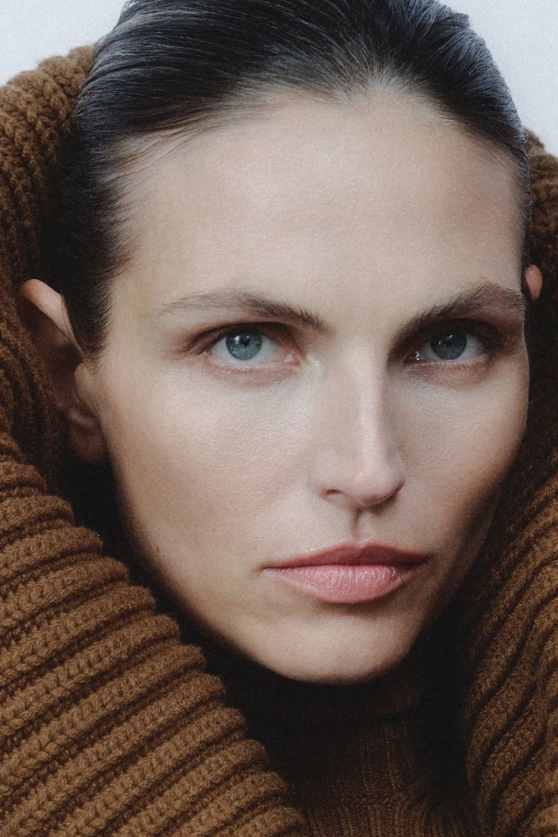 Close-up portrait of a woman with light skin and blue eyes wearing a brown knit turtleneck sweater, looking directly at the camera.