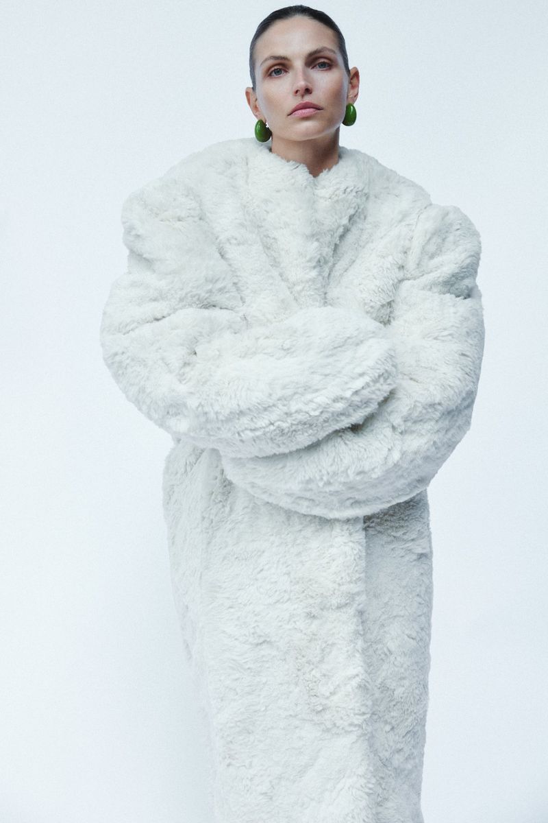 Model Karlina Caune wearing an oversized white faux fur coat and green statement earrings against a pale studio backdrop.