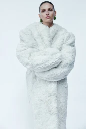 Model Karlina Caune wearing an oversized white faux fur coat and green statement earrings against a pale studio backdrop.