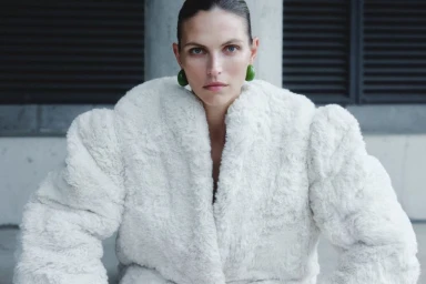 Model Karlina Caune wearing a plush white faux-fur coat and green earrings in an editorial portrait.