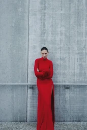 Model wearing a long red dress with a high leg slit leaning against a grey concrete wall