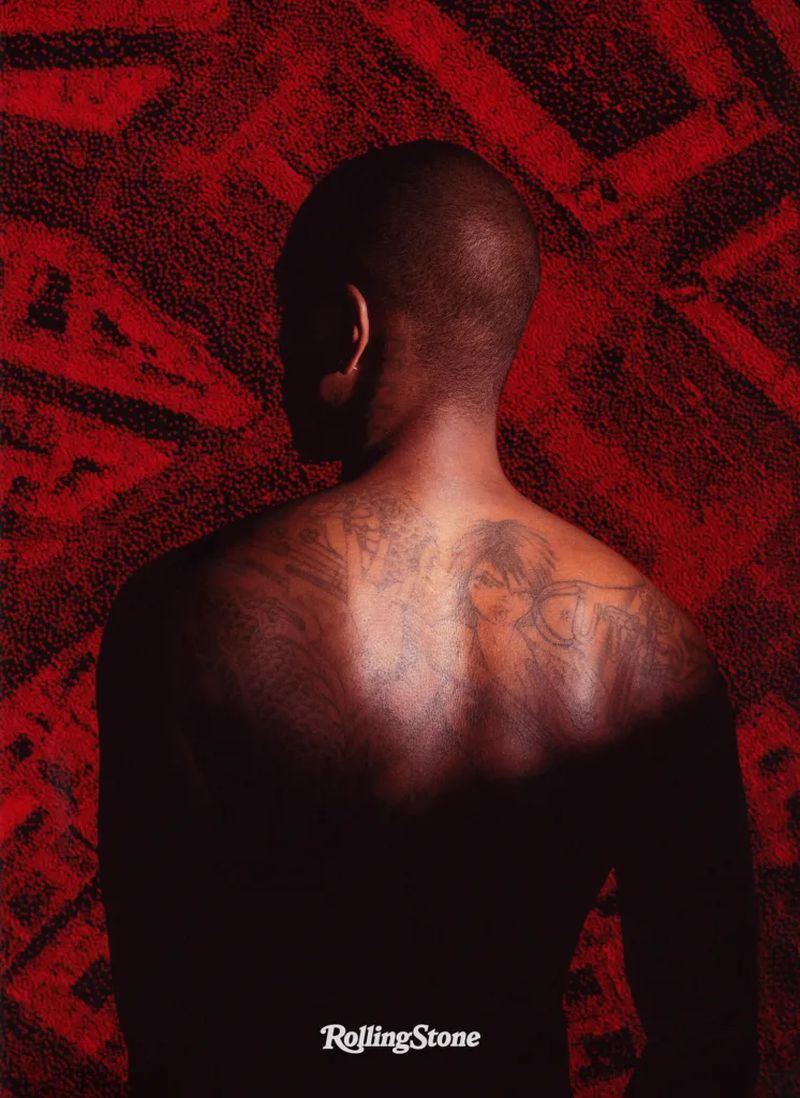 Adult male poses shirtless from the back, showcasing tattoo art, against a textured red backdrop with dramatic lighting in a portrait.