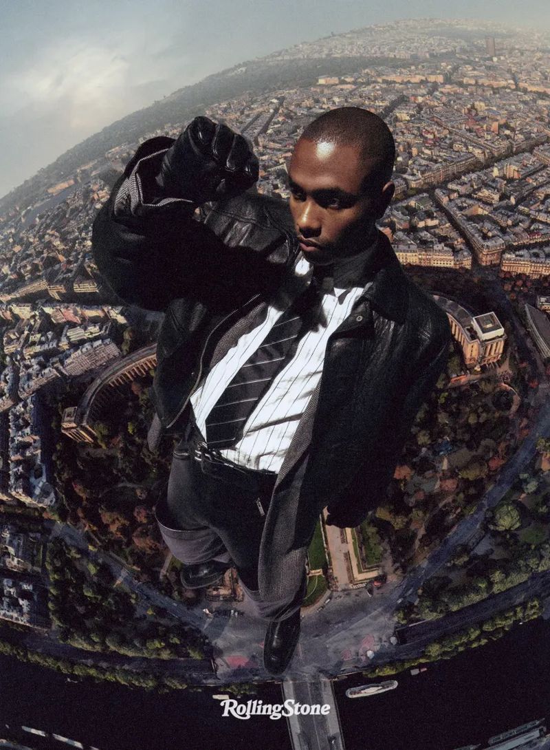 Stylized portrait of a person with slicked-back hair, wearing a black leather jacket and tie, overlaid on a bird's-eye view of a city.