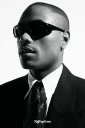 Musician Steve Lacy in dark sunglasses, a black suit jacket, and a white collared shirt with a tie against a plain white background.