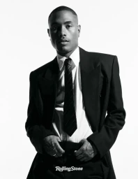 Musician Steve Lacy in a black suit jacket, white collared shirt, and dark necktie against a bright white studio backdrop.