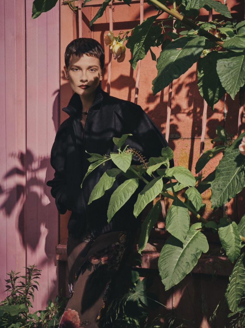 Model Sophie Dominique Gonzalez wearing a black jacket among green foliage against a pink wall with cast shadows.
