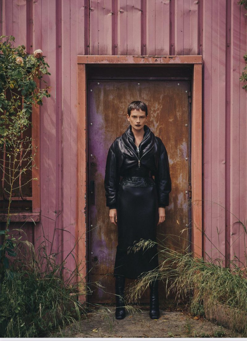 A woman with short hair stands in front of a metal door, wearing a black puffer jacket and skirt, with wild plants in the foreground.