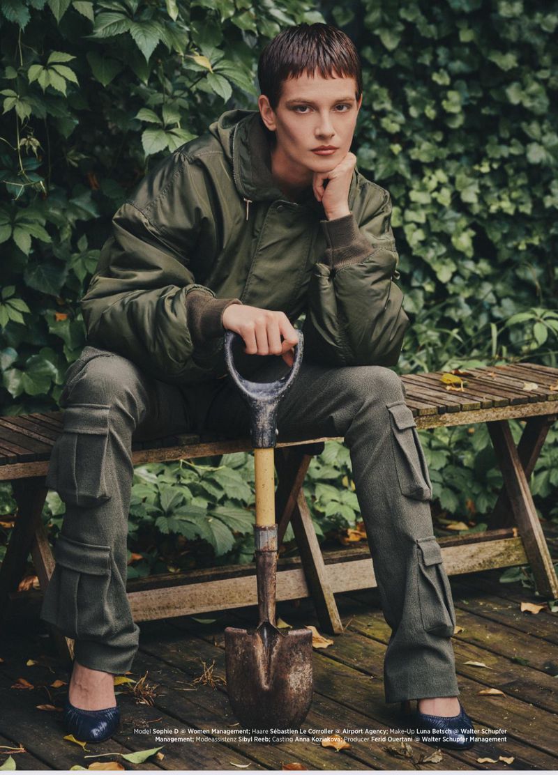 Woman in green bomber jacket and cargo pants sits on a wooden bench, holding a shovel in a lush green outdoor setting, fashion editorial.