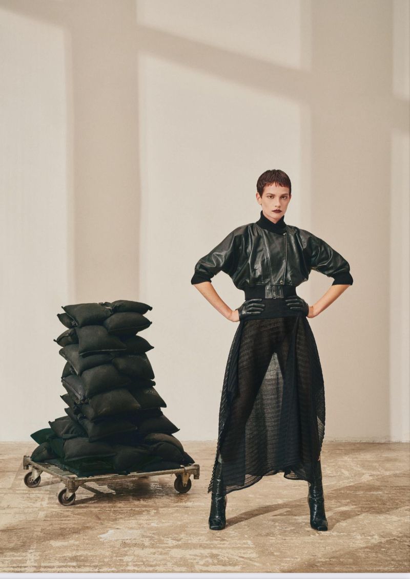 A woman with short brown hair poses indoors with hands on her hips, wearing a black leather jacket, sheer skirt, and boots next to a cart of pillows.
