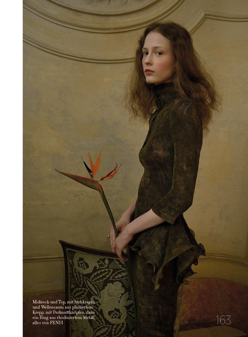 A woman in a green dress stands in front of a wall and vintage chair, holding a bird of paradise flower in a fashion editorial shot.