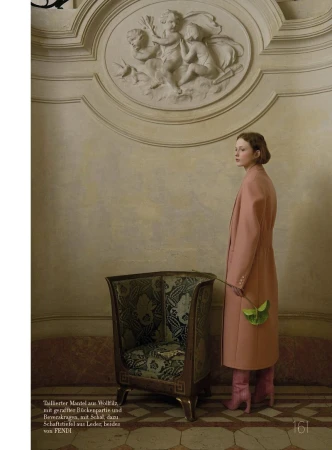 A woman in a pink coat and boots stands in front of a chair with a floral design, in a room with cream walls and marble floors.