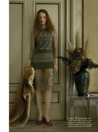A red-haired woman in a lace dress stands in front of a door, holding a fur pelt, next to a vase of dried flowers and a chrome table.