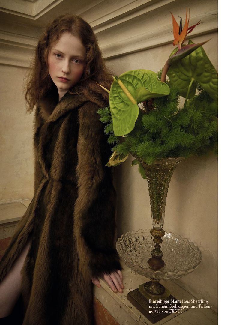 Red-haired woman in a fur coat poses indoors with anthurium and bird of paradise flowers in vases on a marble ledge, fashion shot.