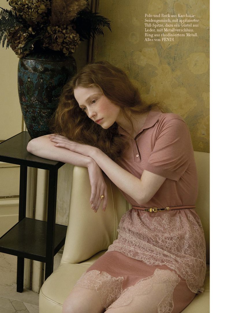 A red-haired woman in a pink dress sits in an indoor setting next to a vase and a small table, creating an elegant and thoughtful composition.