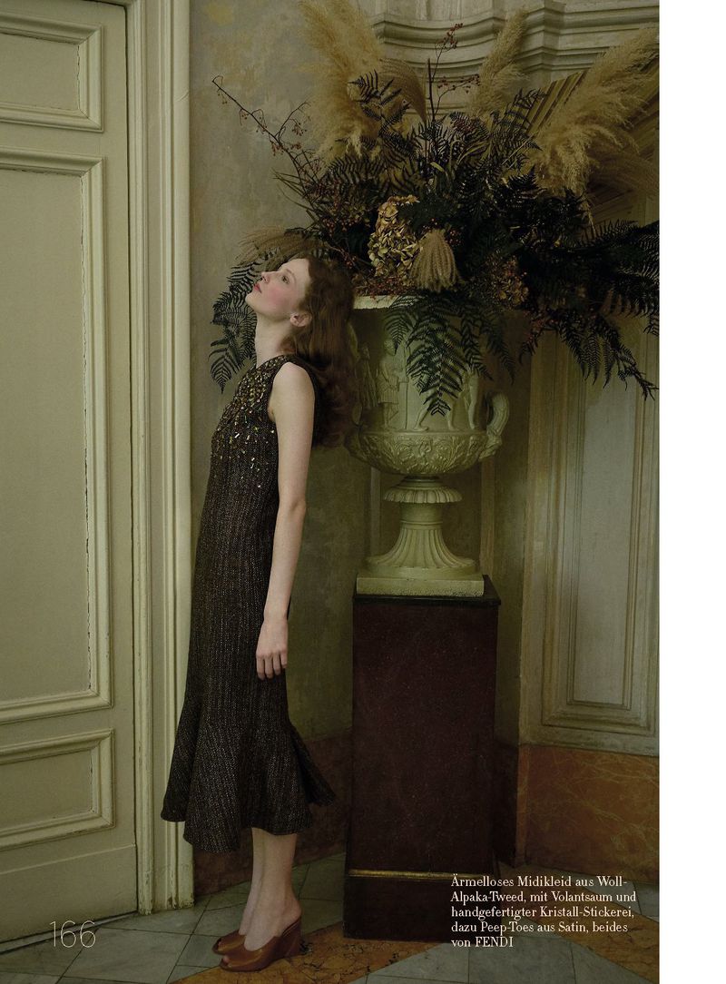 Woman in elegant sleeveless midi dress with embellishments poses next to large urn with floral arrangement in bright indoor space.