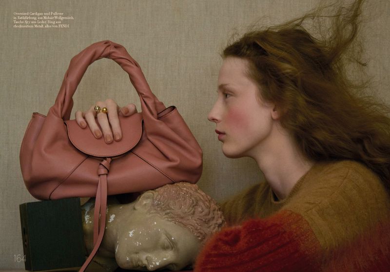 Fashion editorial shot of an adult woman with long brown hair, posed in profile with a dusty rose leather purse and a neutral backdrop.