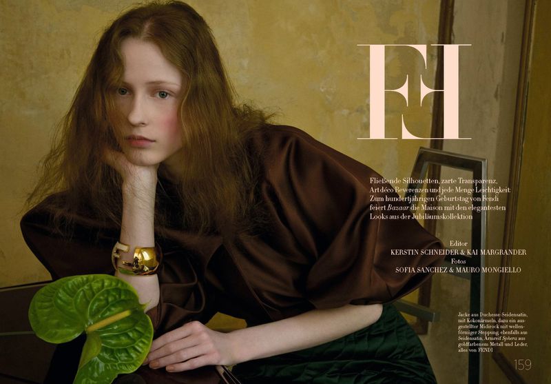 Fashion editorial shot of a young woman with fair skin and auburn hair, wearing a brown satin top and gold bracelet, posing with a plant.