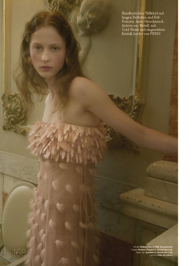 A woman in a light pink feathered dress poses indoors with soft light, a classic bust sculpture, and golden mirror in the background.