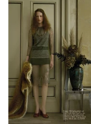 Model standing in front of paneled double doors holding a fur stole next to a tall vase with dried floral arrangements
