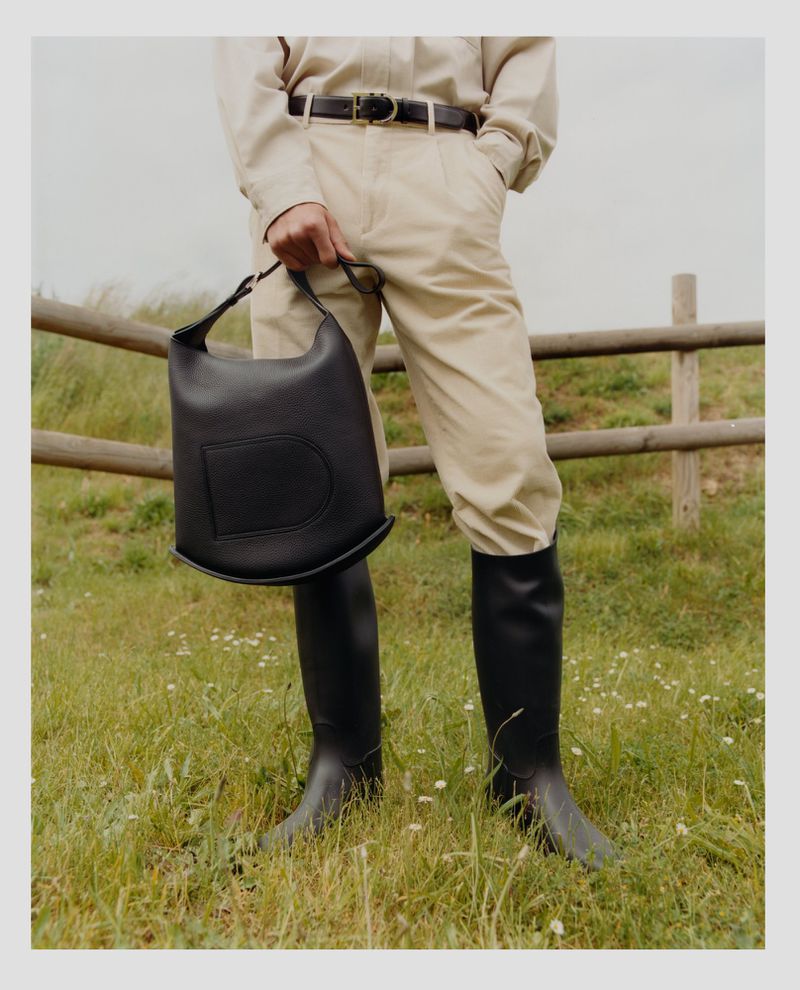 A person stands on a grassy field with a black leather handbag, wearing a tan jumpsuit and black riding boots near a wooden fence.