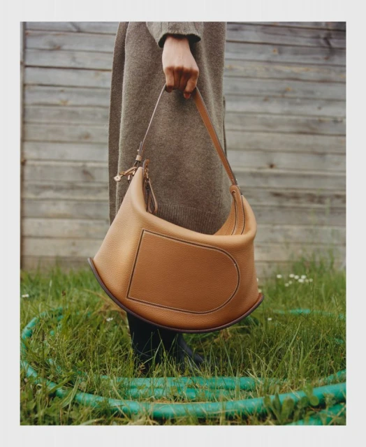 Woman presents a caramel-colored leather shoulder bag with a rectangular pocket outside a weathered wood fence and green grass.