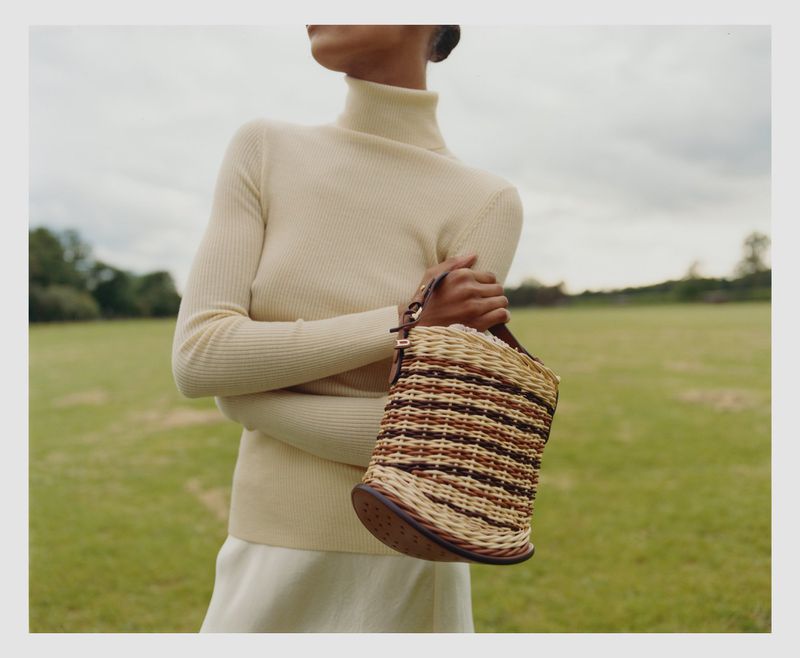 Woman in neutral turtleneck sweater holds woven handbag in a field. Fashion shot in a grassy, outdoor setting with natural lighting.