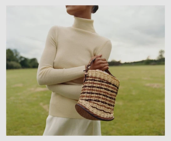 Woman in neutral turtleneck sweater holds woven handbag in a field. Fashion shot in a grassy, outdoor setting with natural lighting.