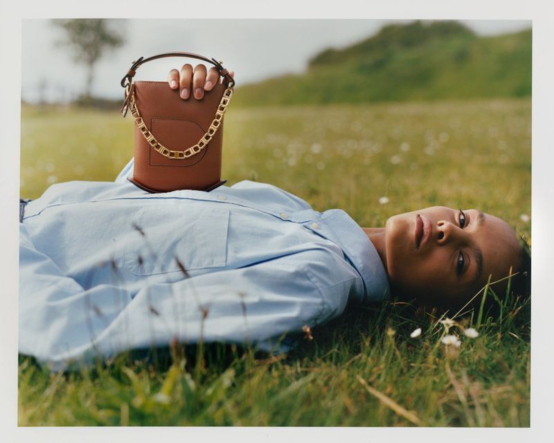 Woman lying on green grass wearing light blue shirt, holding brown leather handbag with gold chain handle outdoors in daylight.