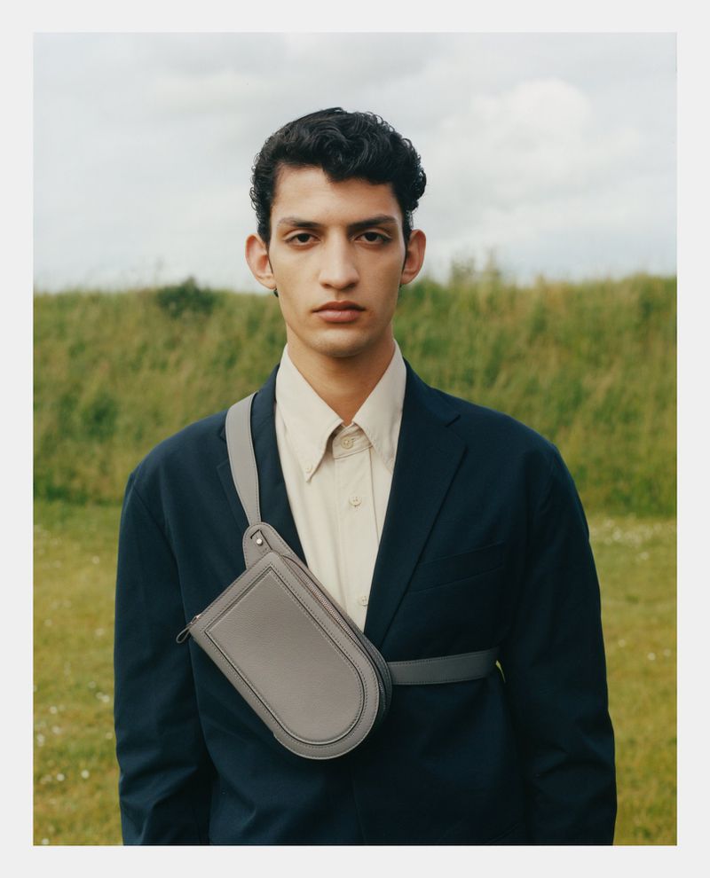 Portrait of a young adult male wearing a blue blazer, light shirt, and gray crossbody bag, posing outdoors in a field with a cloudy sky.