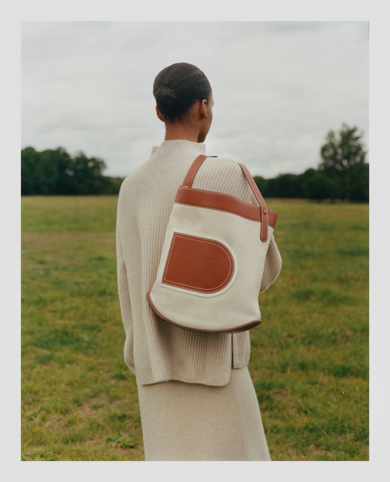 A woman stands in a grassy field wearing a tan sweater and skirt and carrying a cream-colored tote bag with brown leather accents.