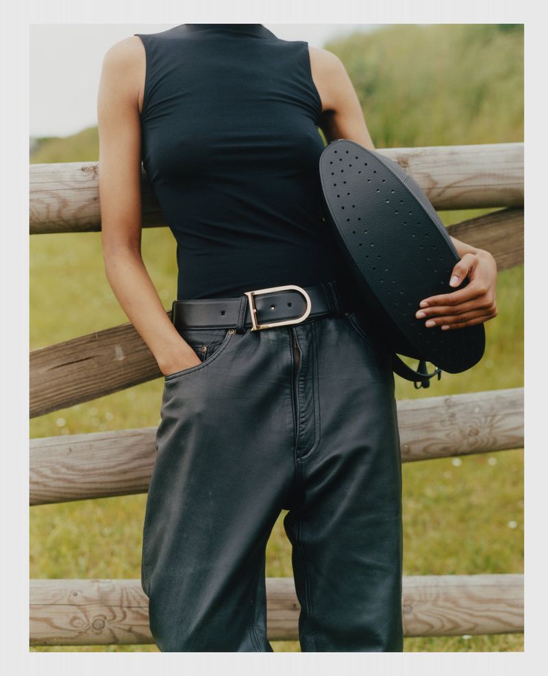 A woman in a black top and leather pants poses with a black helmet against a rural background with a wooden fence.