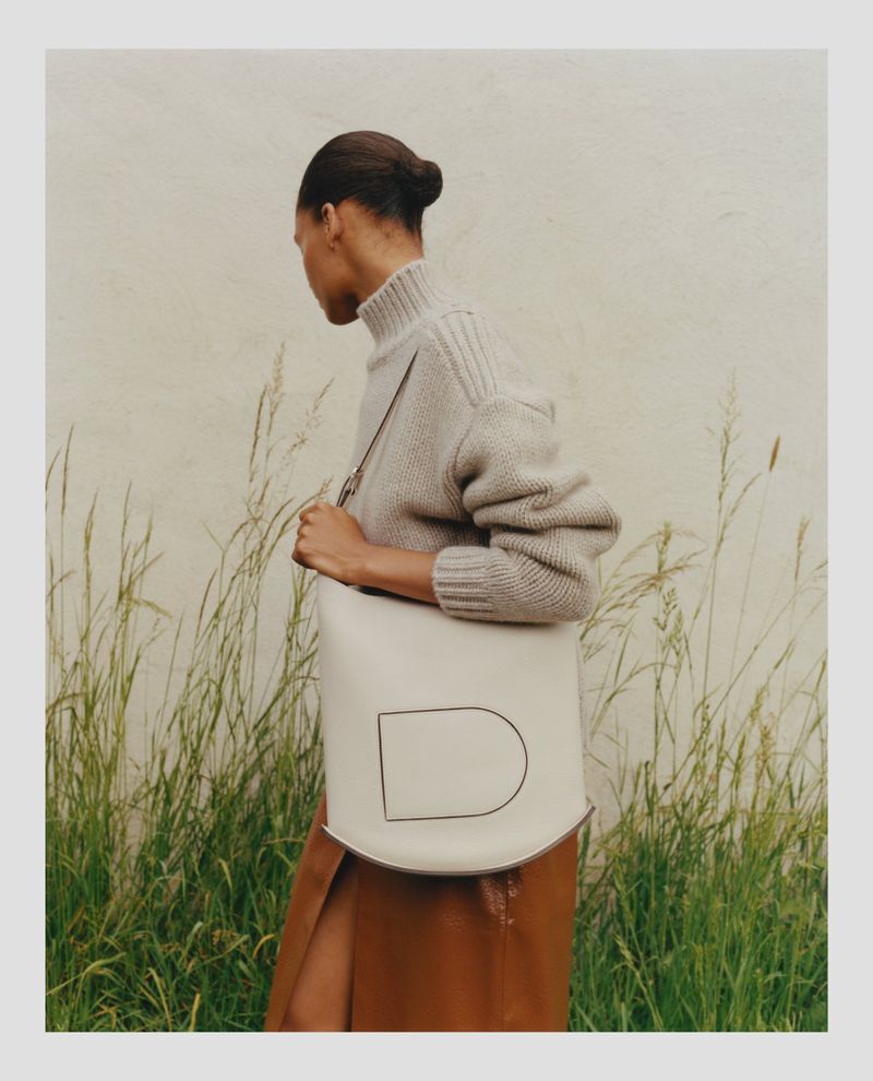 Woman in stylish neutral sweater and brown skirt carrying a cream-colored tote bag with a black line design in an outdoor setting.
