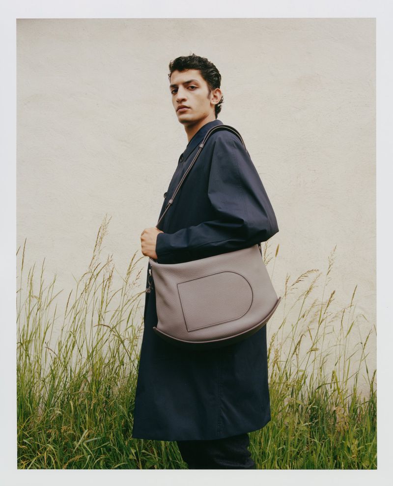 A young adult male model poses with a gray leather messenger bag in front of a grassy field with a light-beige backdrop, fashion photography.