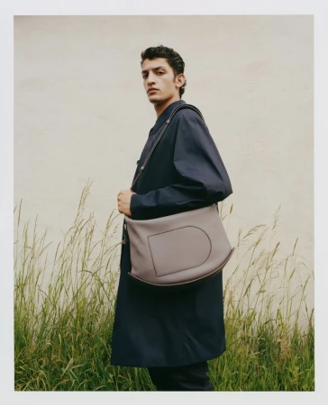 A young adult male model poses with a gray leather messenger bag in front of a grassy field with a light-beige backdrop, fashion photography.