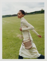 Model in cream turtleneck and matching skirt walking across a green meadow carrying a woven leather shoulder bag.