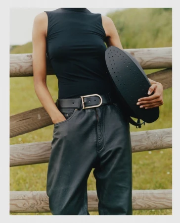 Model wearing a black sleeveless top and dark leather trousers holding a black handbag against a wooden fence