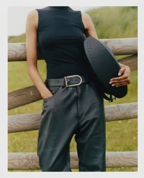 Model wearing a black sleeveless top and dark leather trousers holding a black handbag against a wooden fence