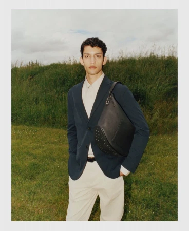 A male model wearing a dark blazer over a light shirt and pale trousers standing outdoors with a leather shoulder bag.
