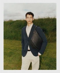 A male model wearing a dark blazer over a light shirt and pale trousers standing outdoors with a leather shoulder bag.