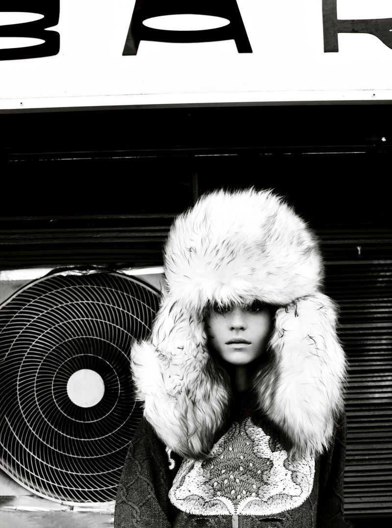 Black-and-white portrait of a model in a fur ushanka hat, posing in front of an air conditioning unit with the text "BAR" above her head.
