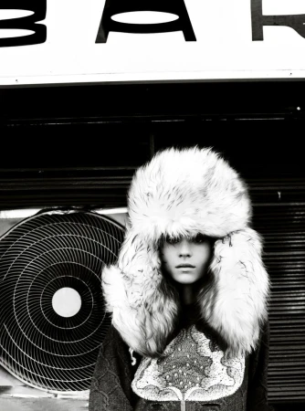 Black-and-white portrait of a model in a fur ushanka hat, posing in front of an air conditioning unit with the text "BAR" above her head.