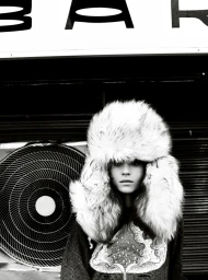 Model wearing a large fluffy faux fur hat and patterned sweater standing in front of a circular fan and a sign reading BAR.